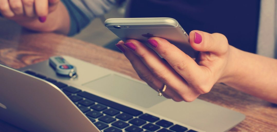 Ein weibliche Hand hält ein Smartphone während die Person am Laptop sitzt